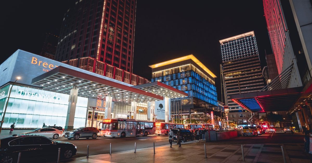 Town with illuminated buildings and skyscrapers near road with cars and buses near pavement with people under dark cloudless sky at night