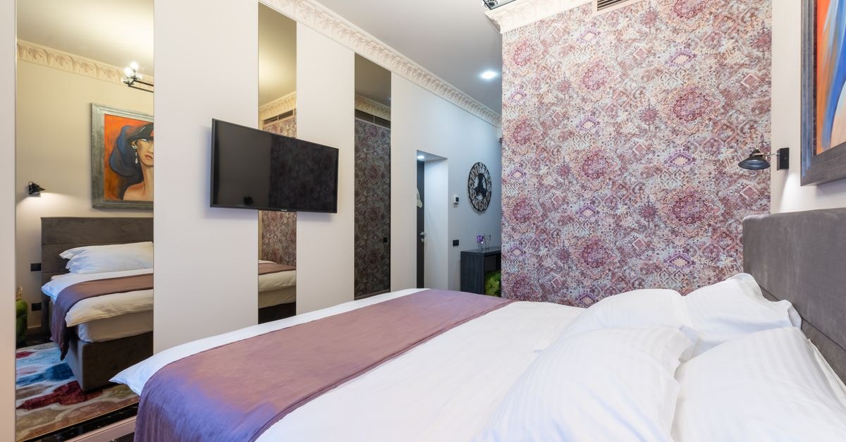 Stylish cozy light hotel suite with bed against TV hanging on wall with mirrors and wall with pink floral ornament