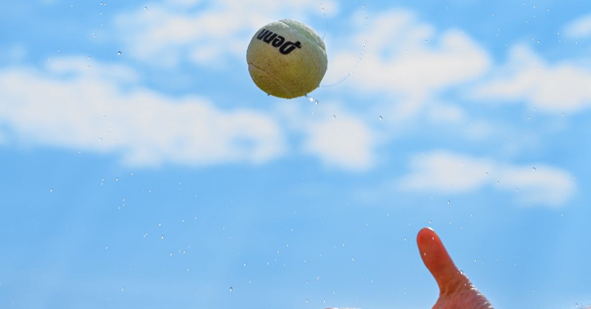 A tennis ball in motion captured mid-air against a clear blue sky with a hand reaching out.