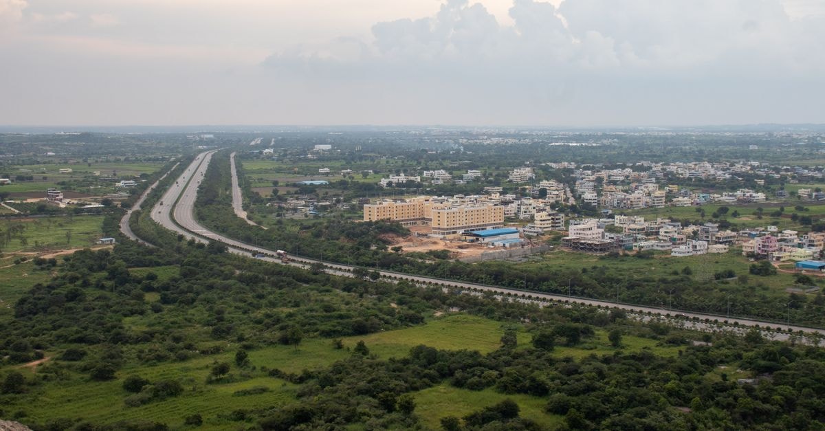 Aerial view of a lush green Indian landscape with a highway stretching through fields and residential areas.