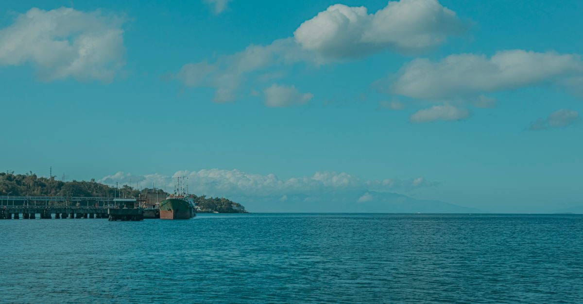 A serene coastal scene with a pier and moored ship under a bright blue sky.
