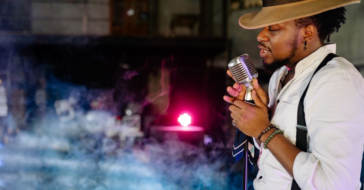 A male singer passionately performs indoors with a vintage microphone, wearing a hat and suspenders.