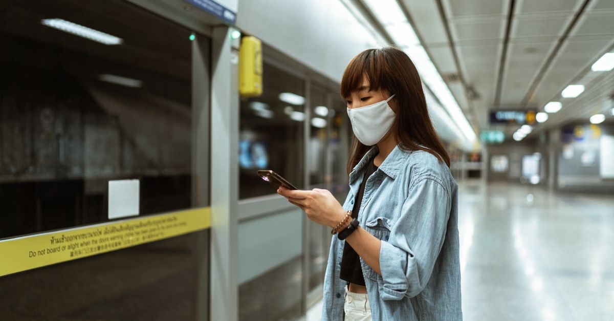 Asian woman wearing a face mask, using her smartphone while waiting at an empty metro station.