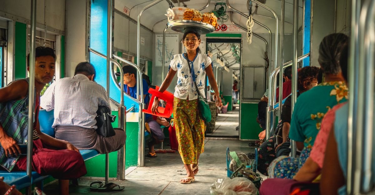 A smiling street vendor walks through a Yangon train with food tray on head.