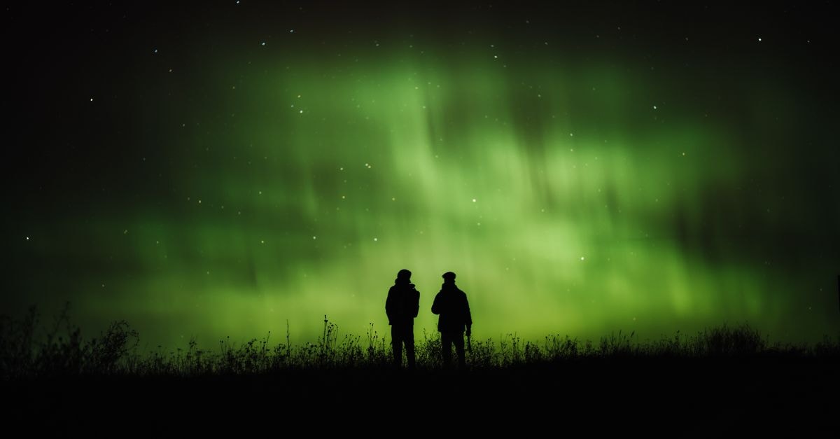 Two silhouetted figures stand beneath the Northern Lights in Kirov Oblast, Russia, creating a serene, dramatic scene.