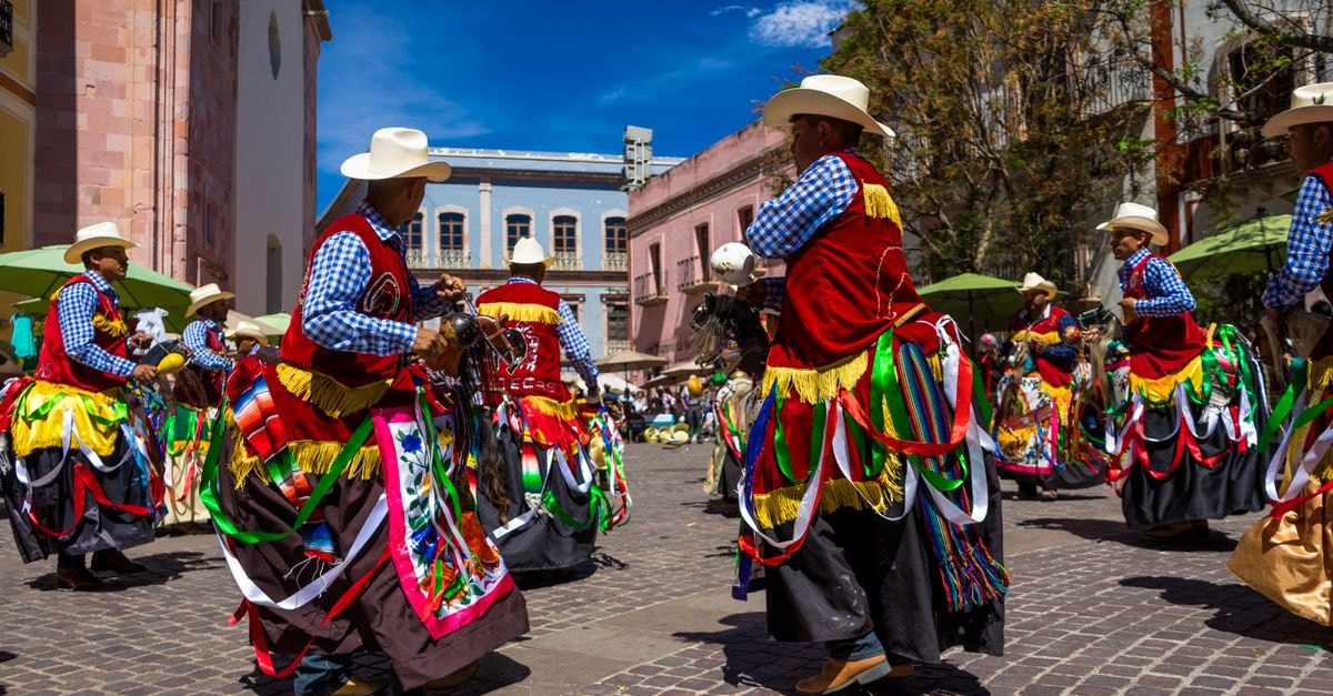 Performers in traditional costumes dance at a lively Mexican street festival under a clear blue sky.