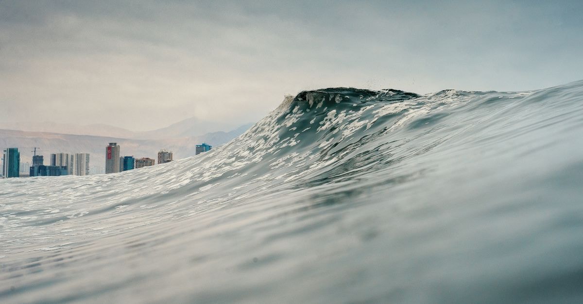 Dramatic ocean wave with Iquique city skyline in the background under a cloudy sky.