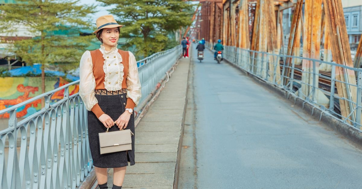 Garco Dragon Hotel in Hanoi: Long Bien Location With Garden and Terrace Spaces Stylish woman standing on an iron bridge with a brown hat and handbag, exuding vintage fashion.