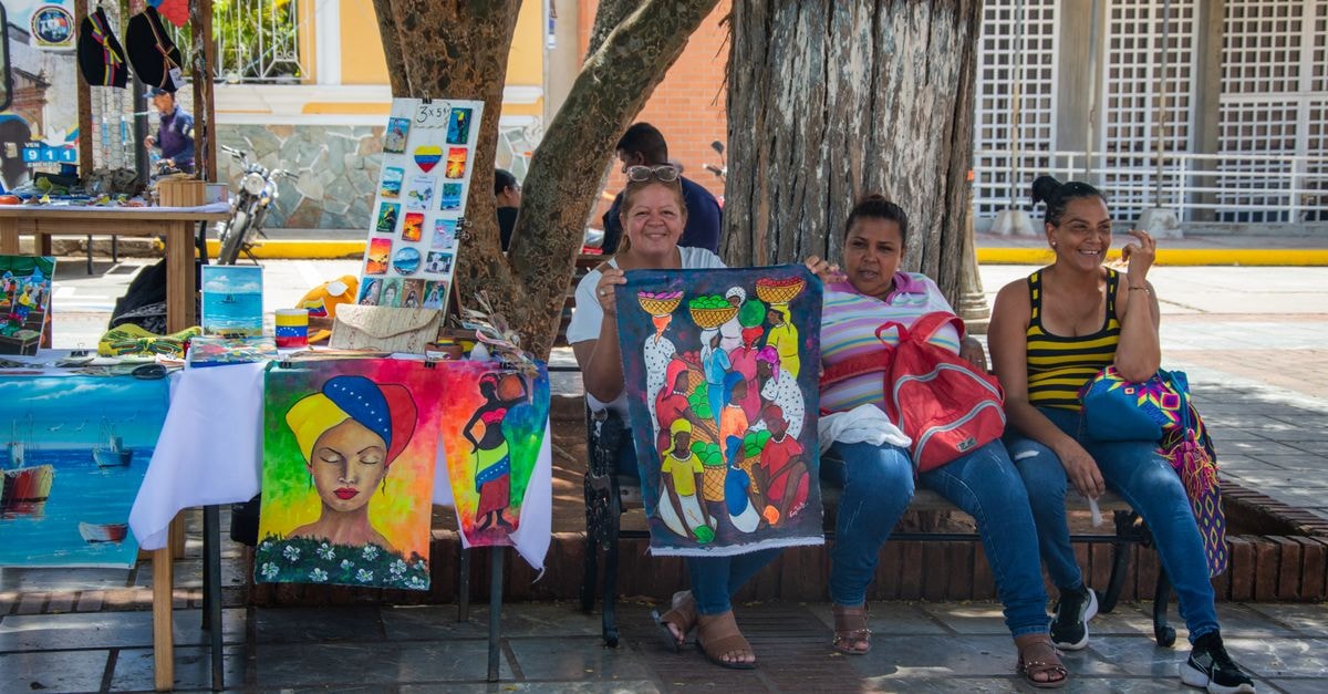 Bria Travel Guide: Art, Culture, and Everyday Flavors Three women sit at a colorful market stall displaying handmade paintings in an urban setting.