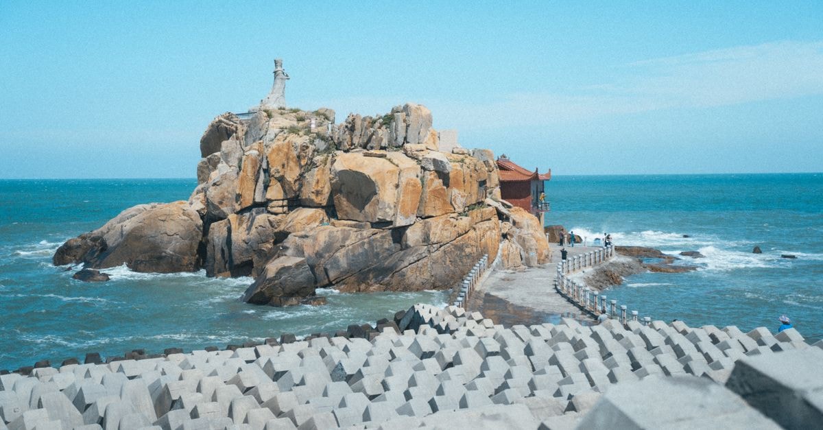 Stunning coastal scene featuring rocky outcrop, ocean and statue under clear blue sky.