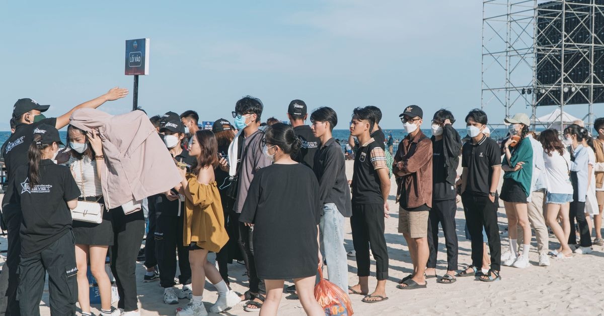 Blackpink's Jennie debuts new single at Lollapalooza, Seventeen's Dino readies alter-ego EP 'Gilboard' A large group of people wearing masks stand in line at a beach event entrance under clear skies.
