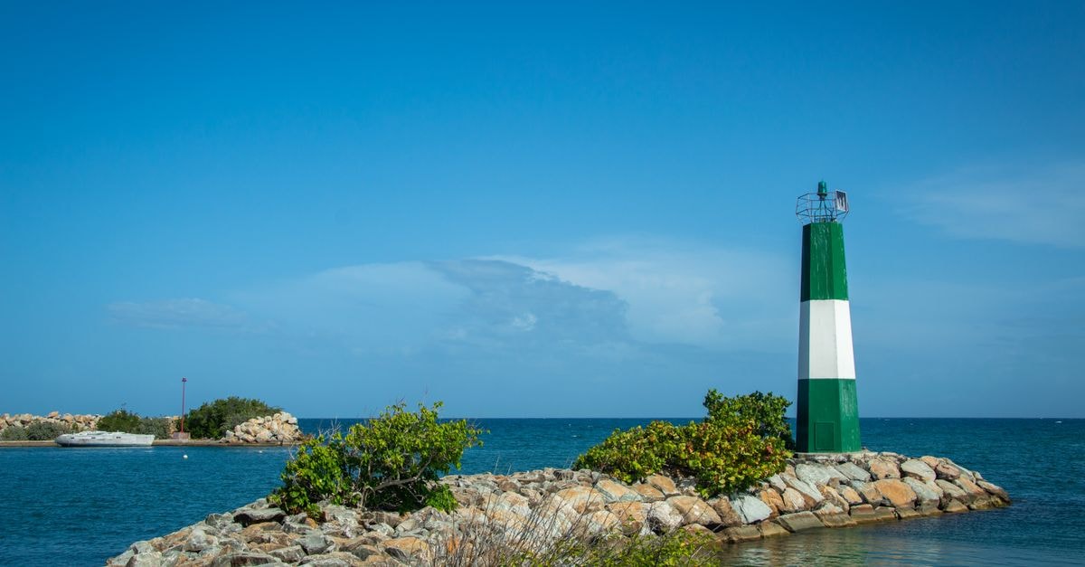 Scenic view of a green and white lighthouse by a calm sea under a clear blue sky.
