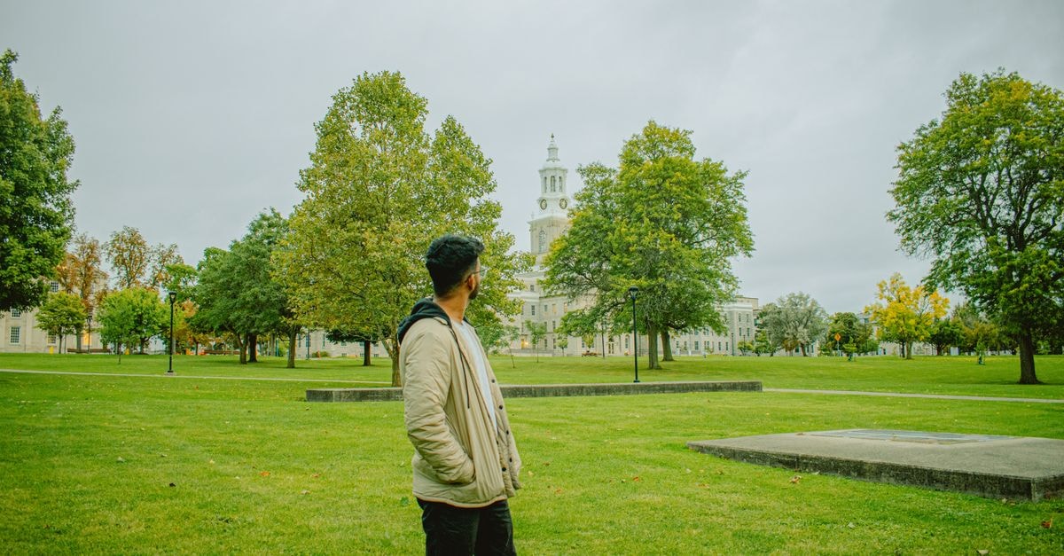A man stands in a park with a historic building and clock tower in the background, surrounded by greenery.