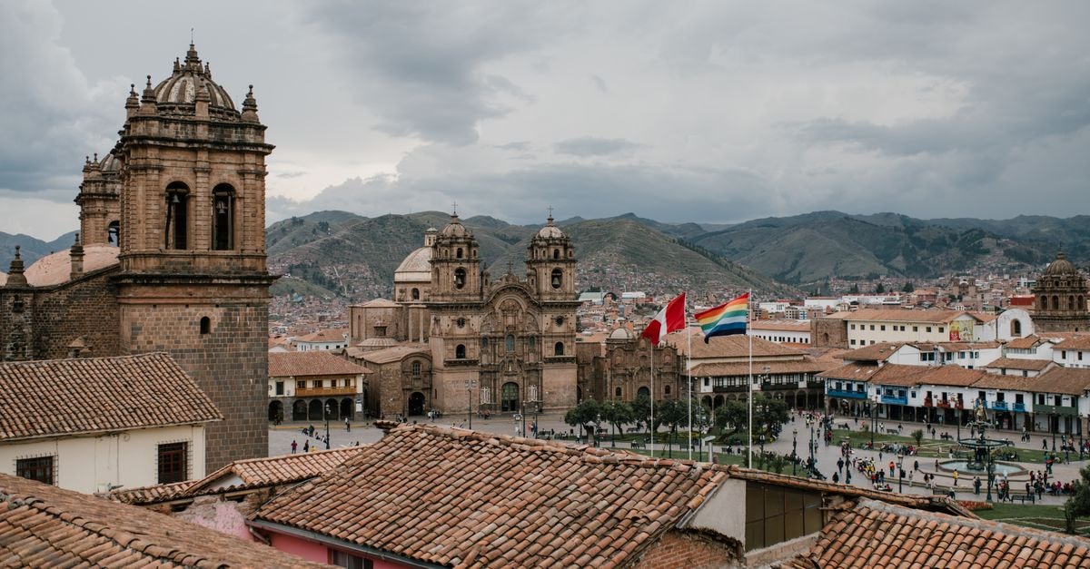Cusco Travel Guide: Planning Tips, Costs and Things to Do High angle of shabby tile roofs of old buildings and Plaza De Armas with national flags and church in Cusco Peru