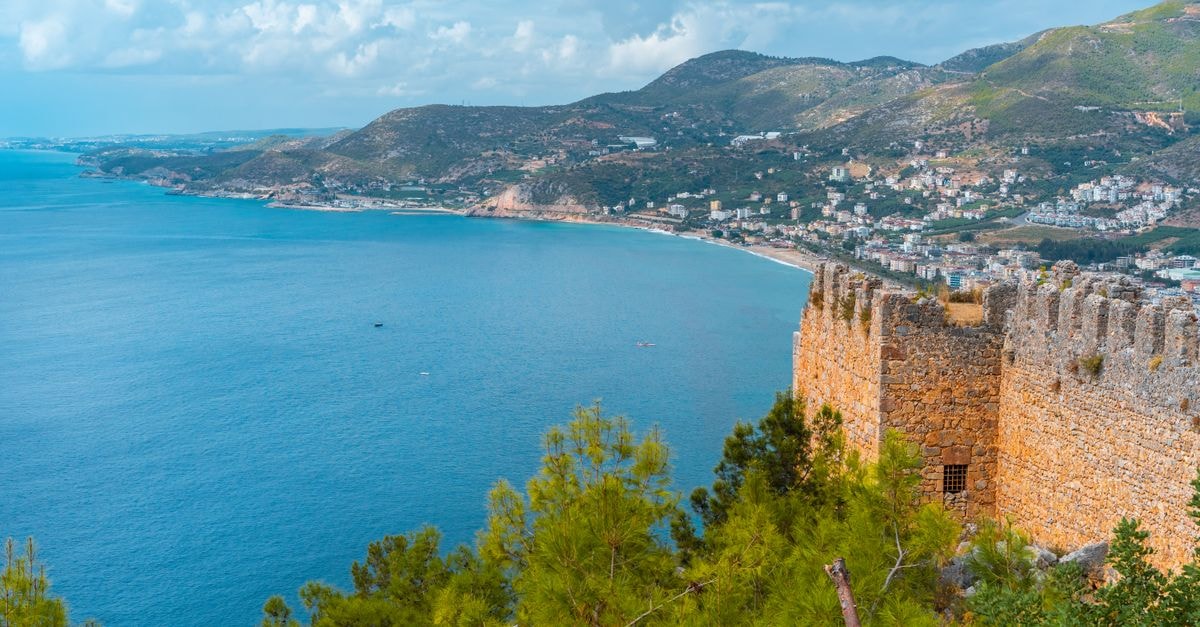 Small village located on grassy hill and weathered castle near sea on coastal area with green trees on summer day