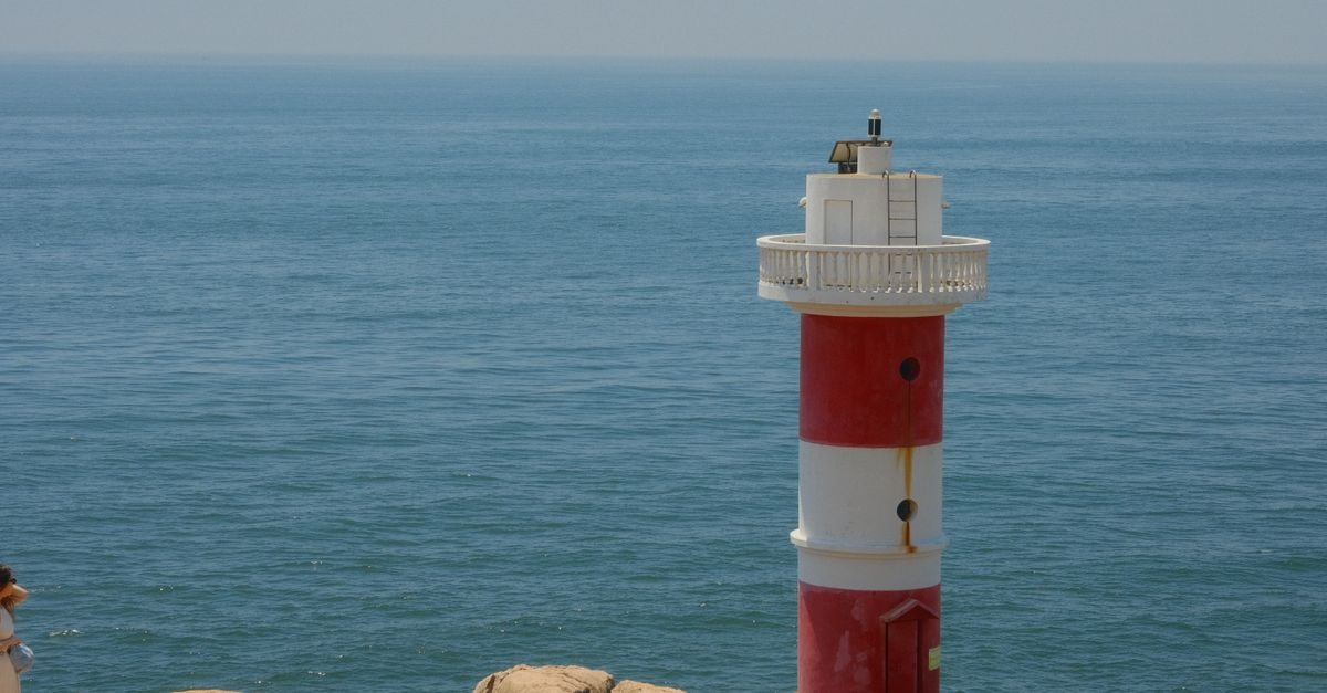 A red and white lighthouse standing on rocky cliffs overlooking the vast ocean under a clear sky.