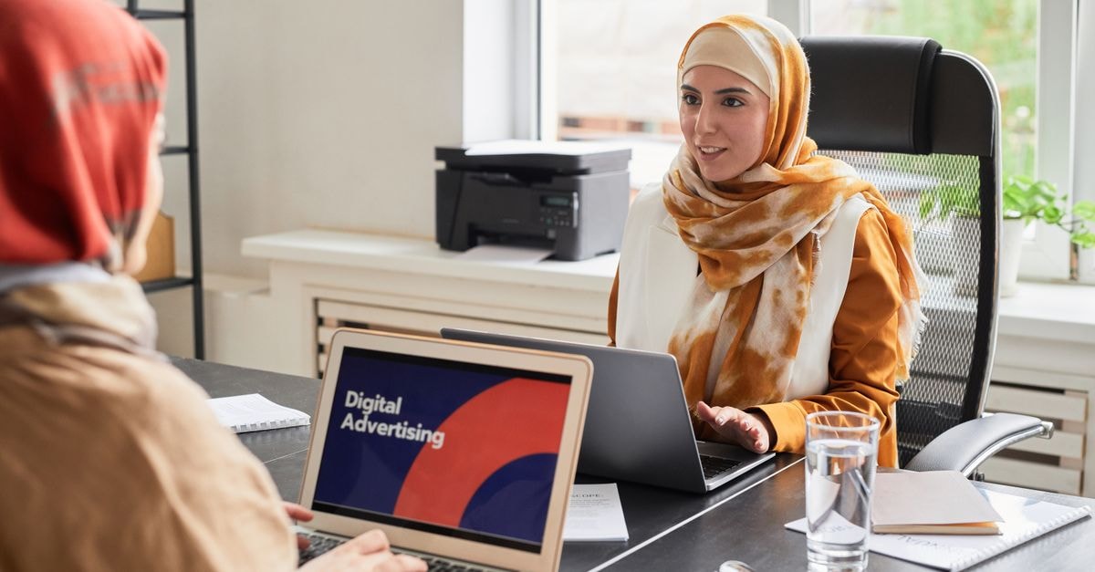 Thryv Holdings: Cash Flow Holds While Revenue Declines Two women in hijabs working on laptops in an office discussing digital advertising.
