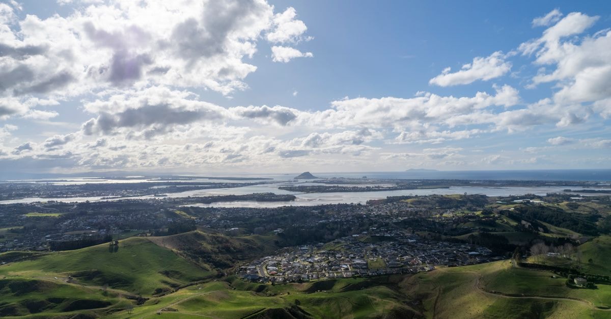 Stunning aerial view of Tauranga, New Zealand with hills, coastline, and clouds under a bright sky.