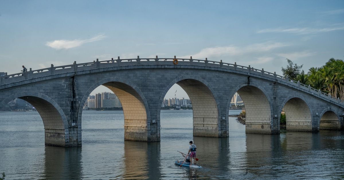 Stone arch bridge spanning a calm river with one person paddleboarding underneath during daytime.