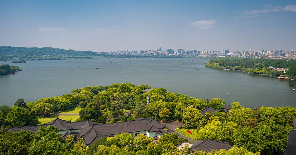 Aerial view over West Lake, lush greenery, and Hangzhou city skyline under a clear blue sky.