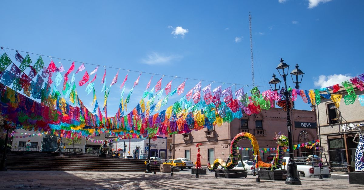Colorful papel picado adorns a plaza in Saltillo, Mexico, under a clear blue sky during a festive celebration.