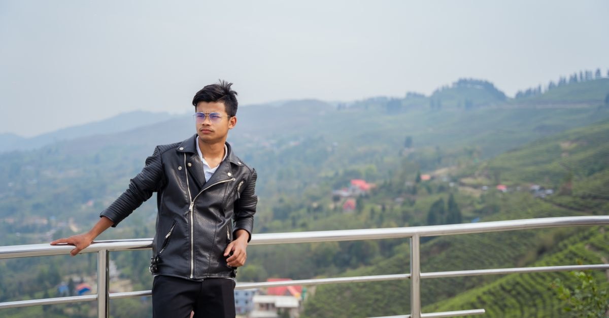 A young man in a leather jacket poses against a scenic mountain view, conveying leisure and style.