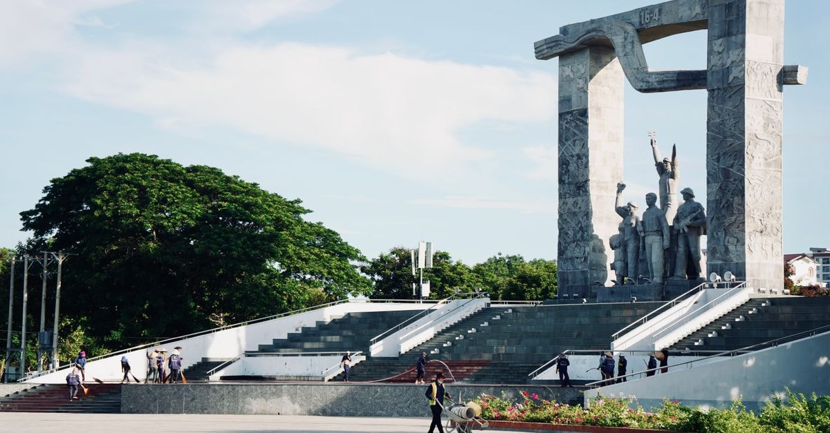 Monumental statue surrounded by greenery in a public park setting with people walking.