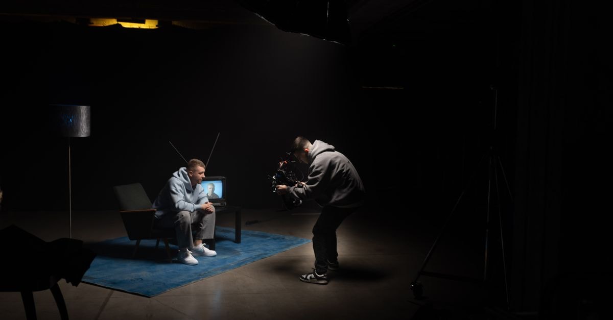 Film production shoot indoors with cinematographer filming an actor sitting, studio lighting.