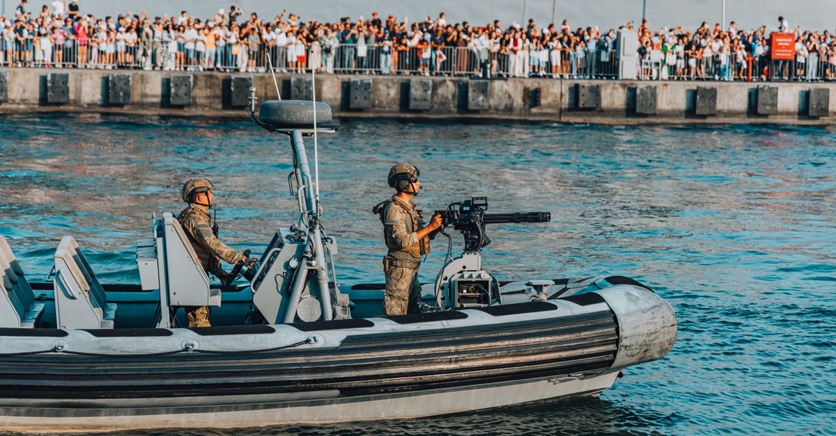 A military boat with armed soldiers patrolling near a crowded pier during a public event.