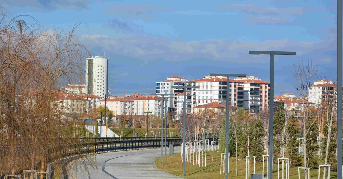 An empty urban park pathway lined with trees, lampposts, and modern residential buildings.