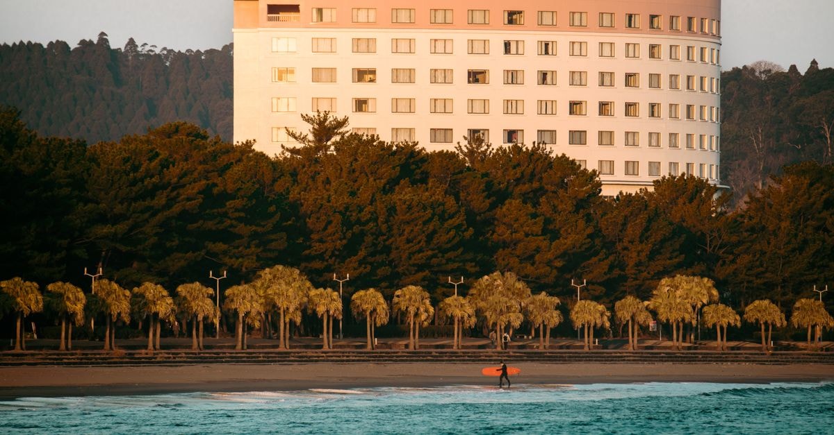 Miyazaki travel guide: a slow, sun-soaked corner of Kyushu Surfer carrying board on Miyazaki Beach with hotel and sunrise backdrop in Japan.