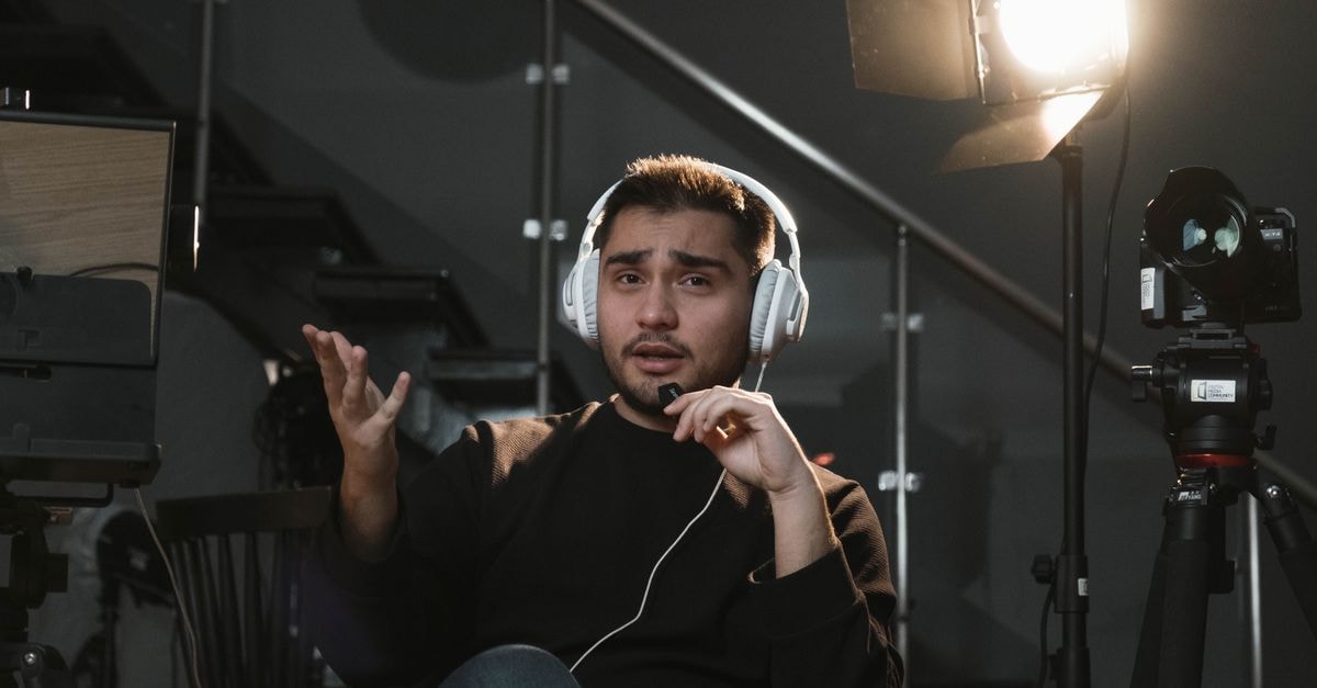 Portrait of a man wearing headphones, sitting in a studio, gesturing towards the camera, with a microphone and lighting setup.