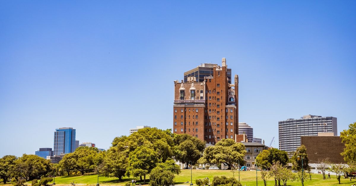 Colonial Lodge Motel in Geelong: Walk to Stadium and Waterfront A tall historic brick building overlooking a park and waterfront under a clear blue sky.