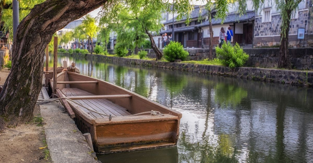 A Slow Travel Guide to Kurashiki's Storied Canals Scenic view of a traditional wooden boat docked along the canal in Kurashiki, Japan, surrounded by lush greenery.