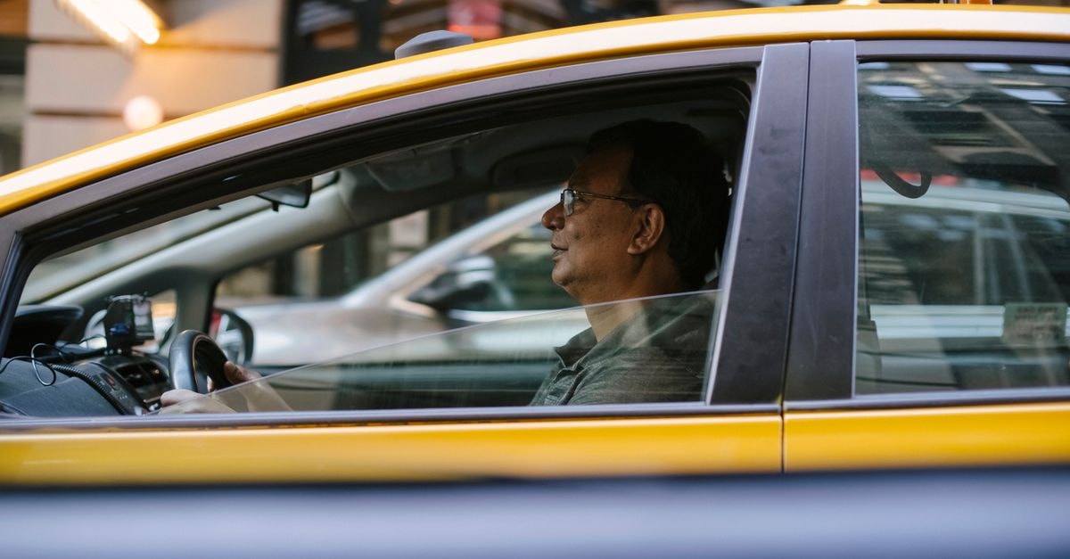 Zoox wins federal approval for paid steering-wheel-free robotaxi rides Side view of ethnic male driver sitting behind steering wheel driving yellow cab with lowered window on city in street with blurred background