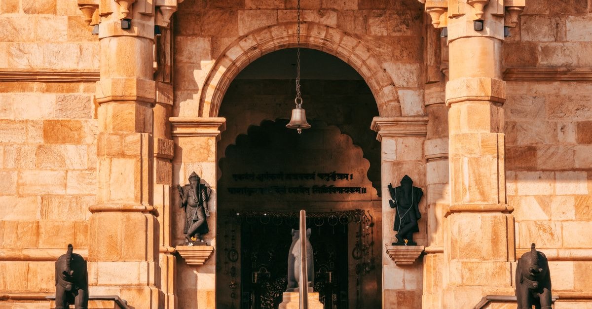 Majestic ancient temple entrance featuring ornate architecture, statues, and a hanging bell in warm sunlight.