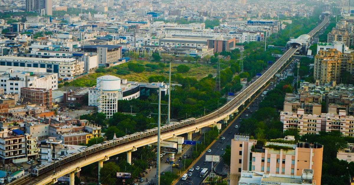 Hotel Rivoli in Rohtak: A First-Class Stay on Sheela Bypass Road Aerial view of a metro train traversing an urban cityscape with dense buildings and greenery.