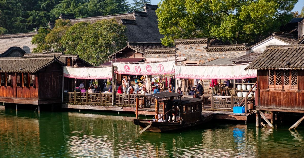 Jinhua Travel Guide: Things to Do in the Heart of Zhejiang Charming riverside market with wooden structures and lush greenery in an Asian village setting.