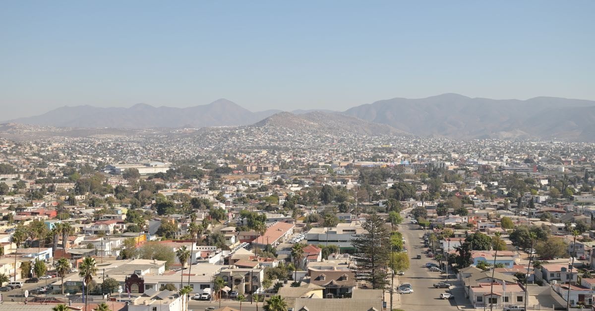 Aerial daytime view of Tijuana's cityscape with mountainous backdrop under a clear sky.