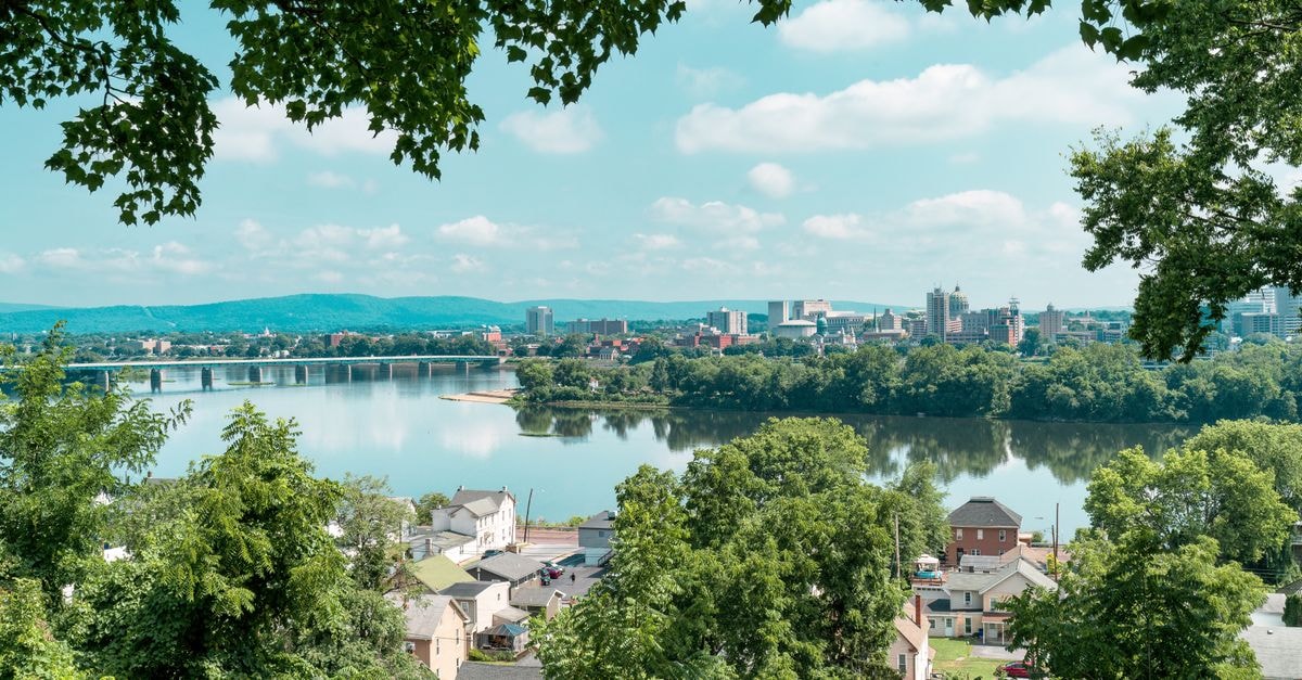 Hilton Harrisburg, Harrisburg — A 1990 Capitol-Area Landmark A picturesque view of a city skyline reflected in a calm river, framed by lush trees.