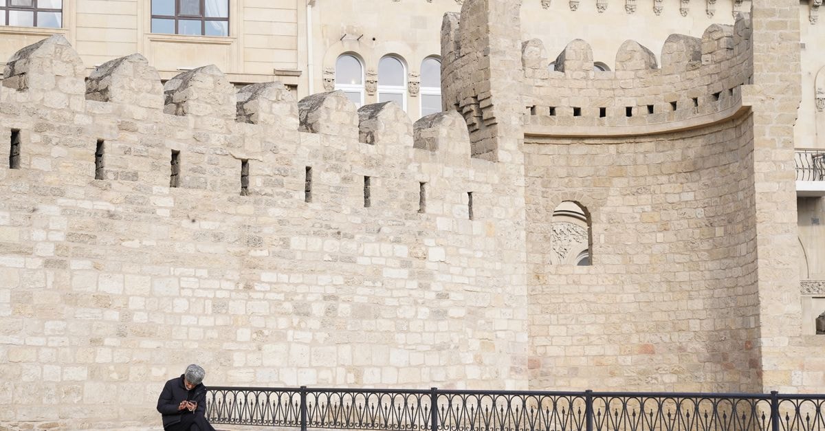 A lone man sits by an ancient stone wall in a historic city setting.