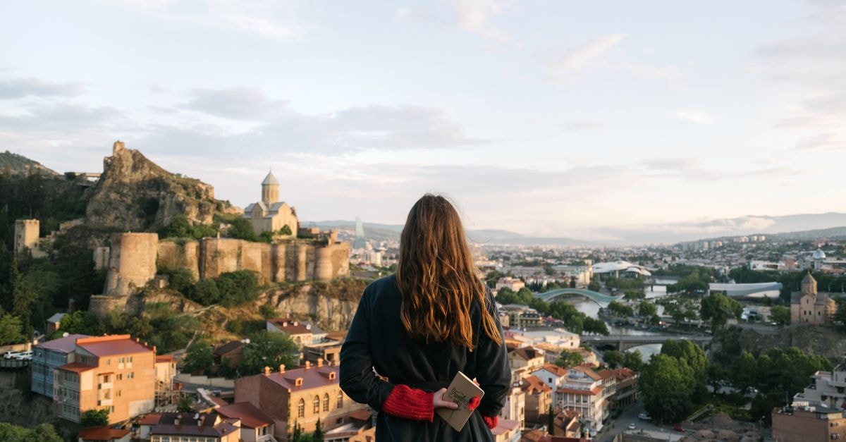 A woman stands with a notebook, gazing at the Tbilisi skyline and historic fortress, capturing a moment of nostalgia.