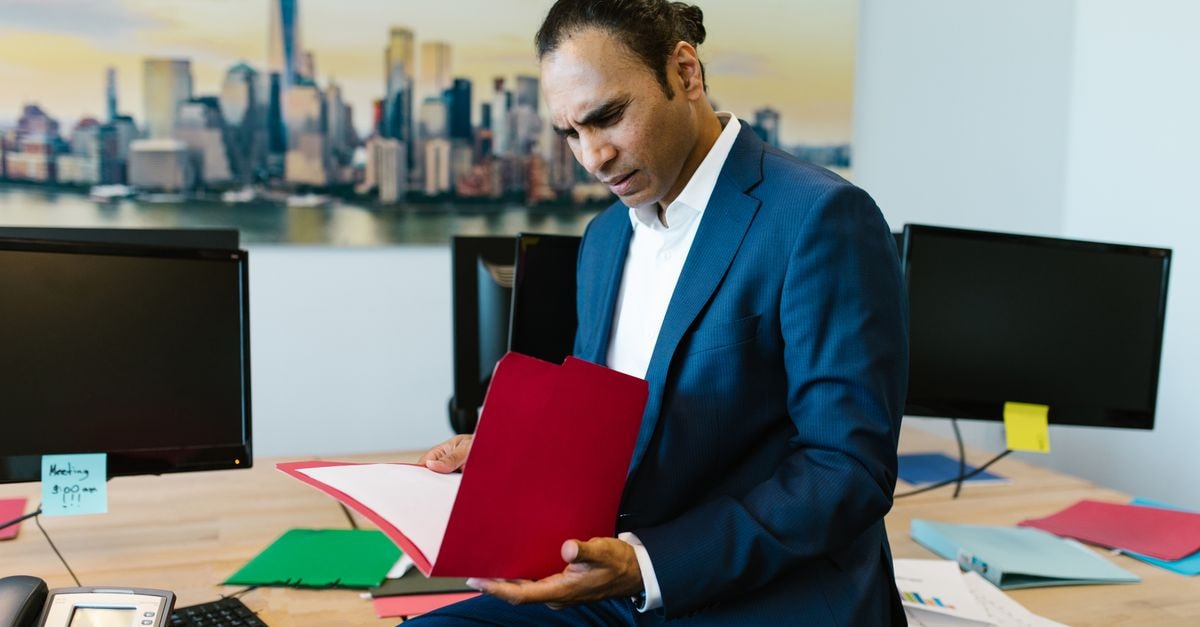 Businessman in an office reviewing documents with a serious expression, surrounded by computers and paperwork.