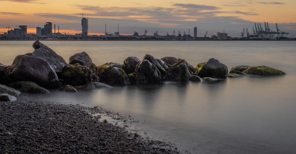 Serene coastal view with industrial skyline at twilight, showcasing calm waters and rocks.