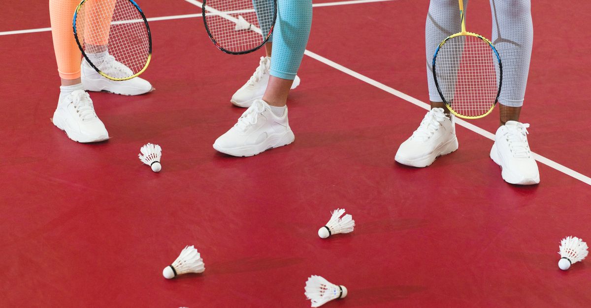 Three badminton players with rackets and shuttlecocks on a red court wearing active wear.