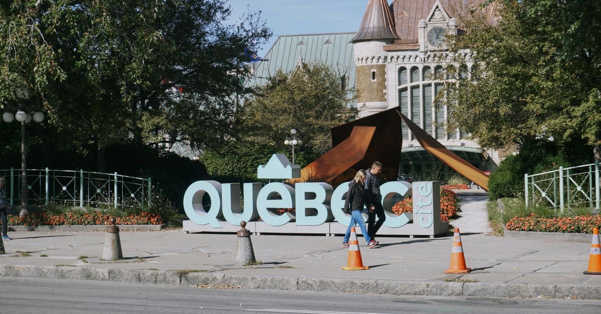 Tourists stroll by the famous Québec City sign in a sunny outdoor setting.