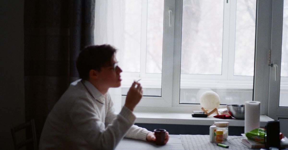 Slow Mornings and Quiet Tables: A Saransk Travel Guide Contemplative young man smoking indoors beside a bright window. Soft focus enhances the calm mood.