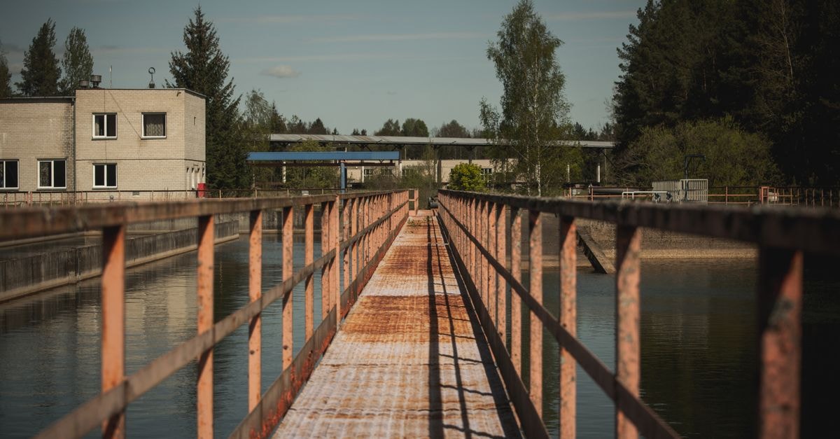 A Quiet Border Crossing: A Travel Guide to Valga, Estonia A rusty pedestrian bridge stretches over water in an industrial area with trees in Lithuania.
