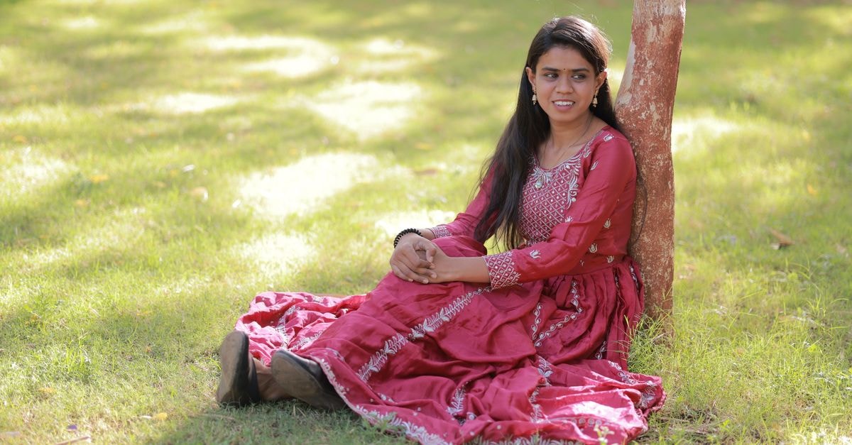 A young woman in a red traditional dress rests against a tree in a sunny park.