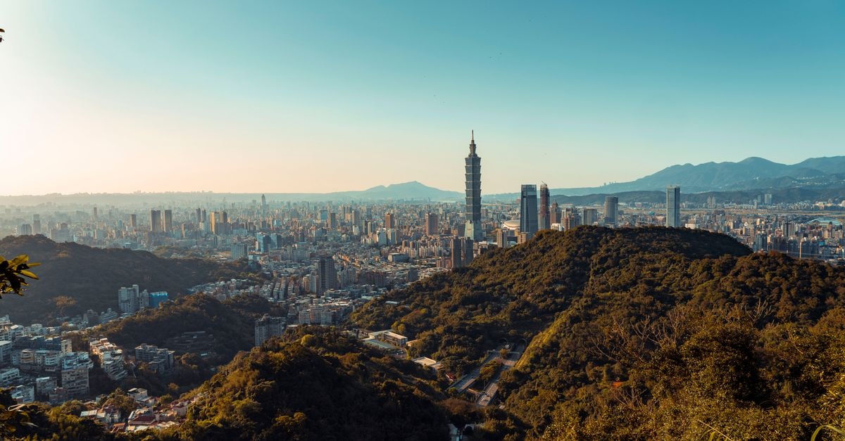 A beautiful cityscape of Taipei including Taipei 101 and lush hills under a clear blue sky.